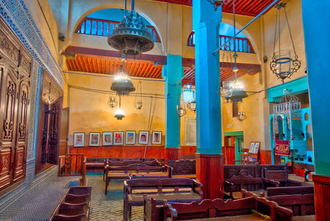 The interior of the Lazama Synagogue in Marrakech — its turquoise columns, hanging bronze lanterns, warm-toned walls and zellige-tiled floor create a colourful and intimate space that has served the Jewish community of Marrakech since it was founded in 1492