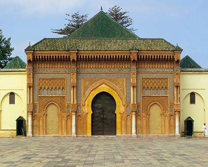 The ornate golden gates of the Royal Palace of Rabat — the brass-studded cedar doors set within a zellige-tiled facade mark the entrance to the Mechouar, the official seat of the Moroccan monarchy, whose grounds remain closed to the public but whose exterior is one of the most photographed facades in the capital