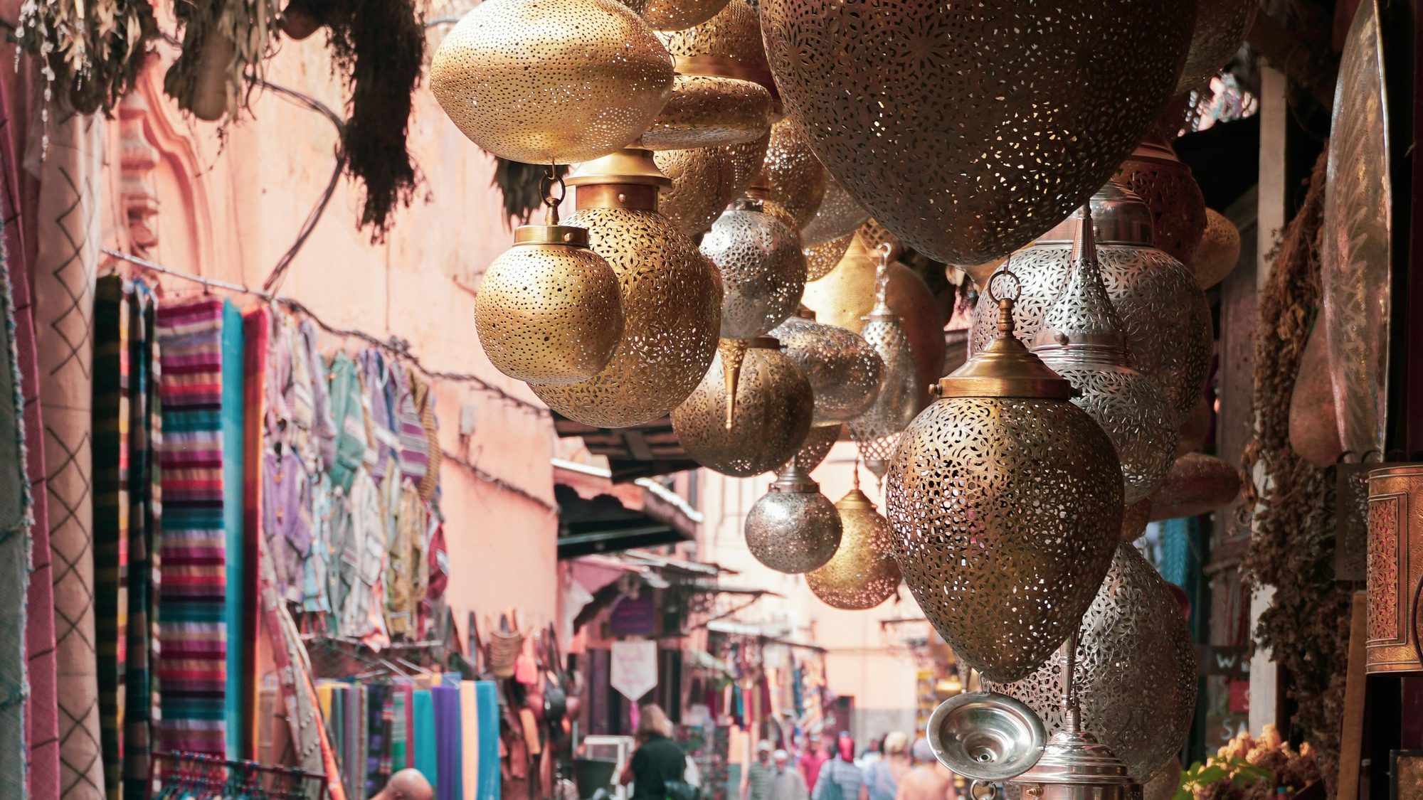 Pierced brass and metal lanterns hanging above a Moroccan souk lane lined with brightly coloured woven textiles
