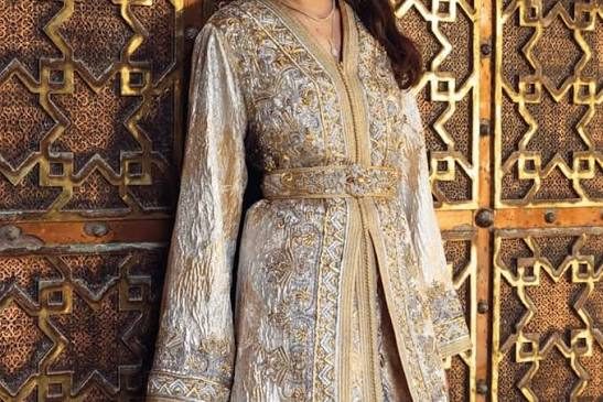 A pale gold caftan with embroidered front panels and a matching belt, worn beside a carved wooden door