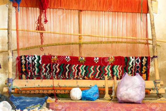 An upright wooden loom with orange warp threads and a colourful woven band in progress, with balls of wool at its base