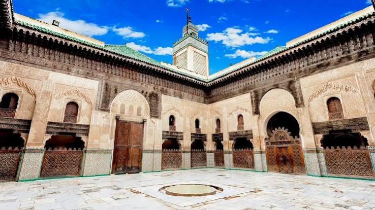 Courtyard of a Marinid-era madrasa in Fez with zellige-covered lower walls, carved plaster, carved cedar upper walls and a minaret beyond