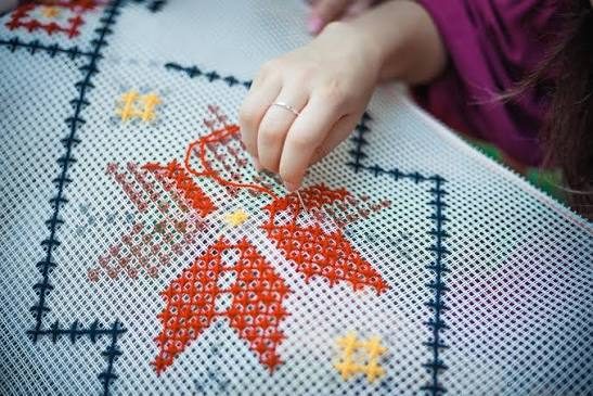 Hands working red counted-thread embroidery onto white even-weave fabric with a blue border