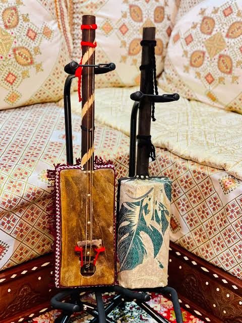 Two guembri stringed instruments with rectangular skin-covered bodies, standing upright on a stand