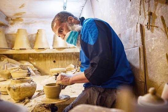 A potter in a blue jacket shaping a vessel on a wheel in a clay-dusted workshop