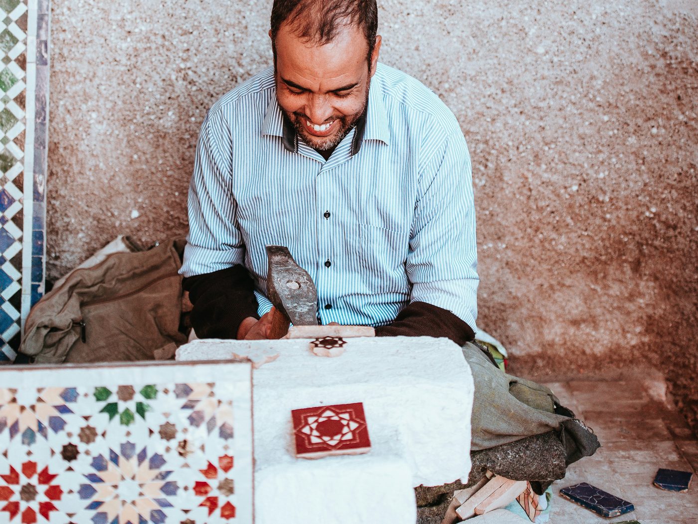 A zellige craftsman smiling as he shapes a glazed tile piece with a hammer on a stone block, a star-pattern panel in front of him