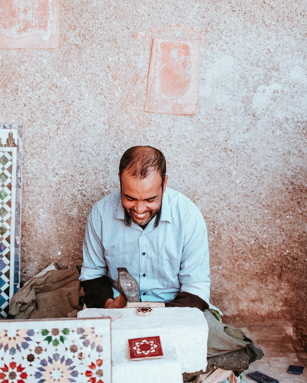 A smiling craftsman seated at a stone block, shaping a glazed tile piece with a hammer, with a finished zellige panel beside him